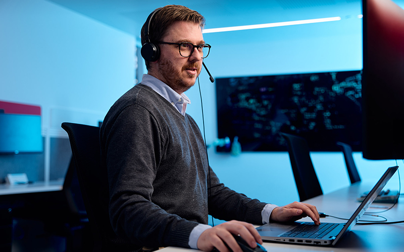 A data analyst studies a dual screen setup with a headset on in an office setting. The background has a light blue hue.
