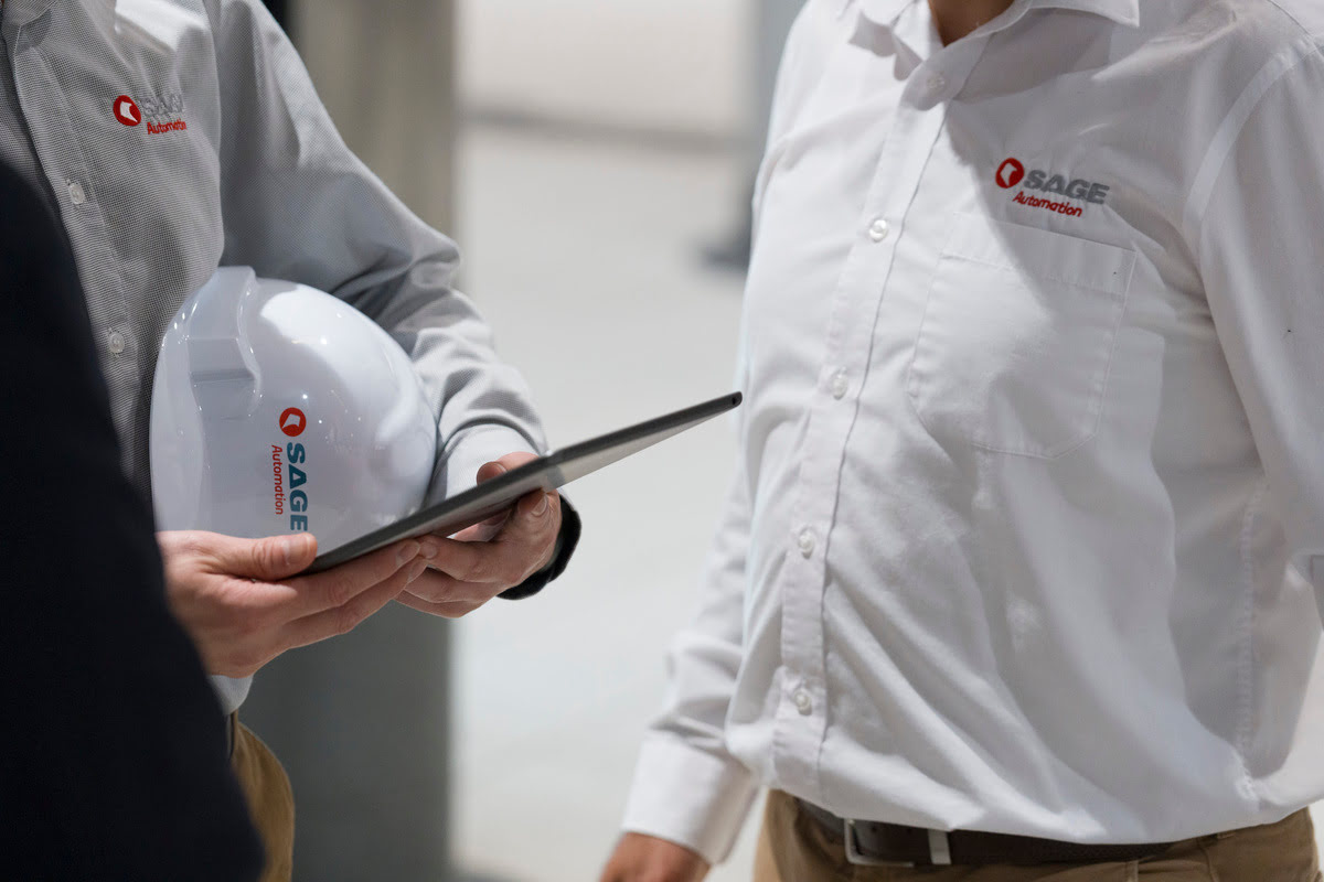 Two SAGE Automation employees in uniform shirts holding a tablet and a white hard hat.